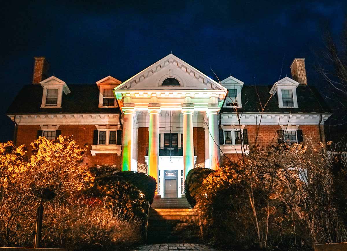 A historic brick mansion illuminated at night, featuring large front columns and surrounded by overgrown vegetation.