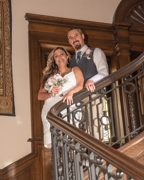 A couple in formal attire poses on a staircase, smiling and holding flowers.