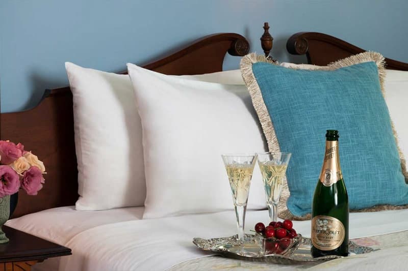 A neatly made bed with white pillows and a blue cushion, featuring two glasses of champagne and a bottle on a tray with cherries.