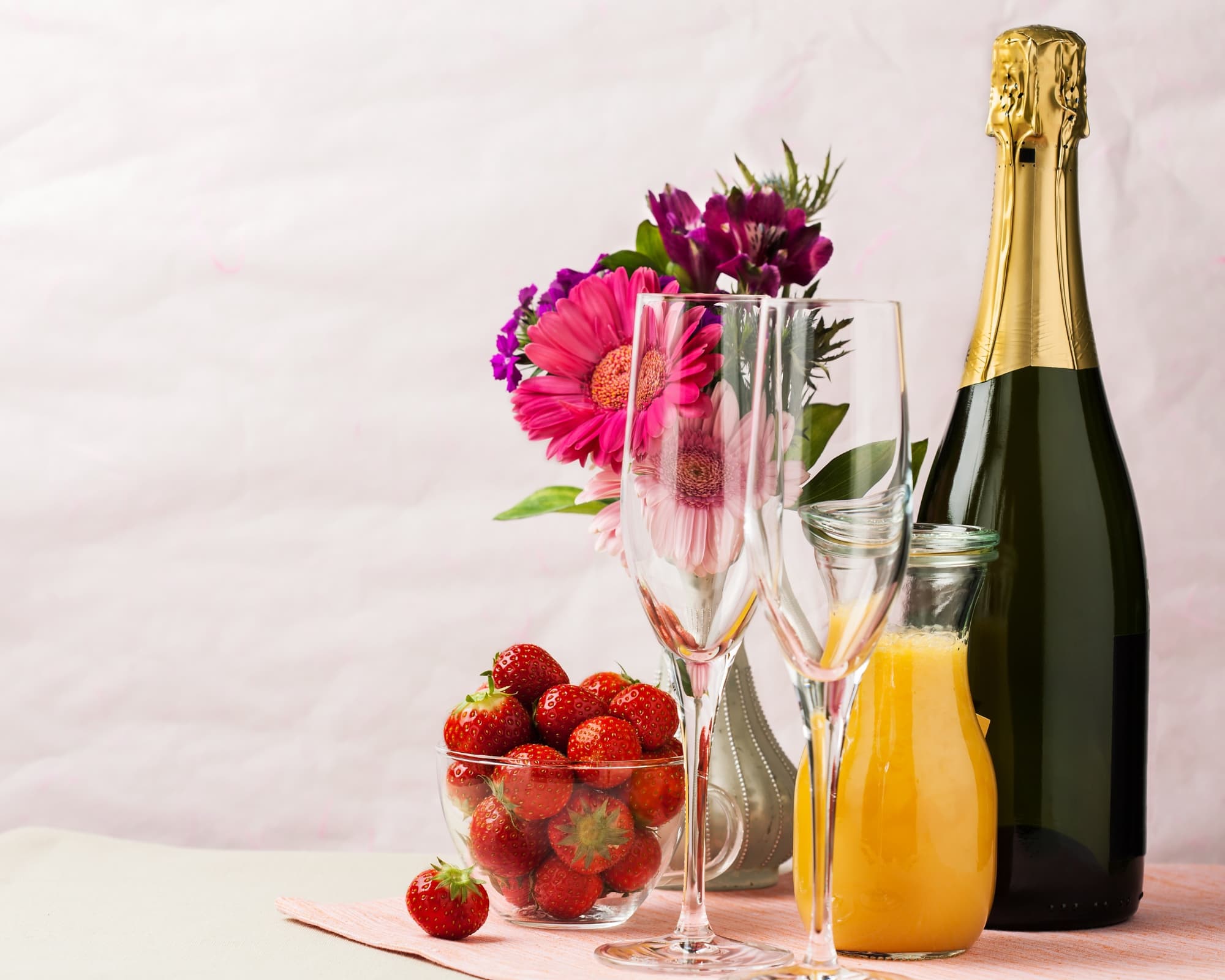 A bottle of champagne, two empty glasses, a bowl of strawberries, orange juice, and colorful flowers are arranged on a table.