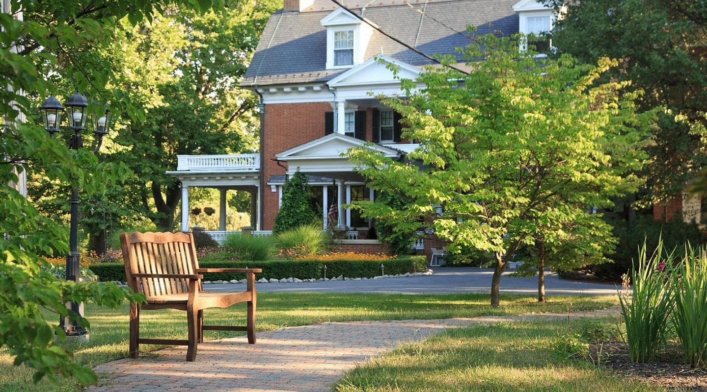 A wooden bench sits in a path leading to a picturesque brick house surrounded by greenery.