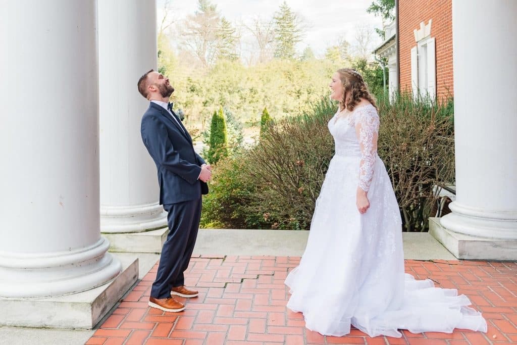 A bride in a white wedding dress stands facing a groom in a dark suit beneath tall columns.