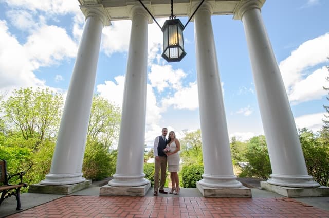 A couple stands together under a large lantern and tall columns with greenery in the background.