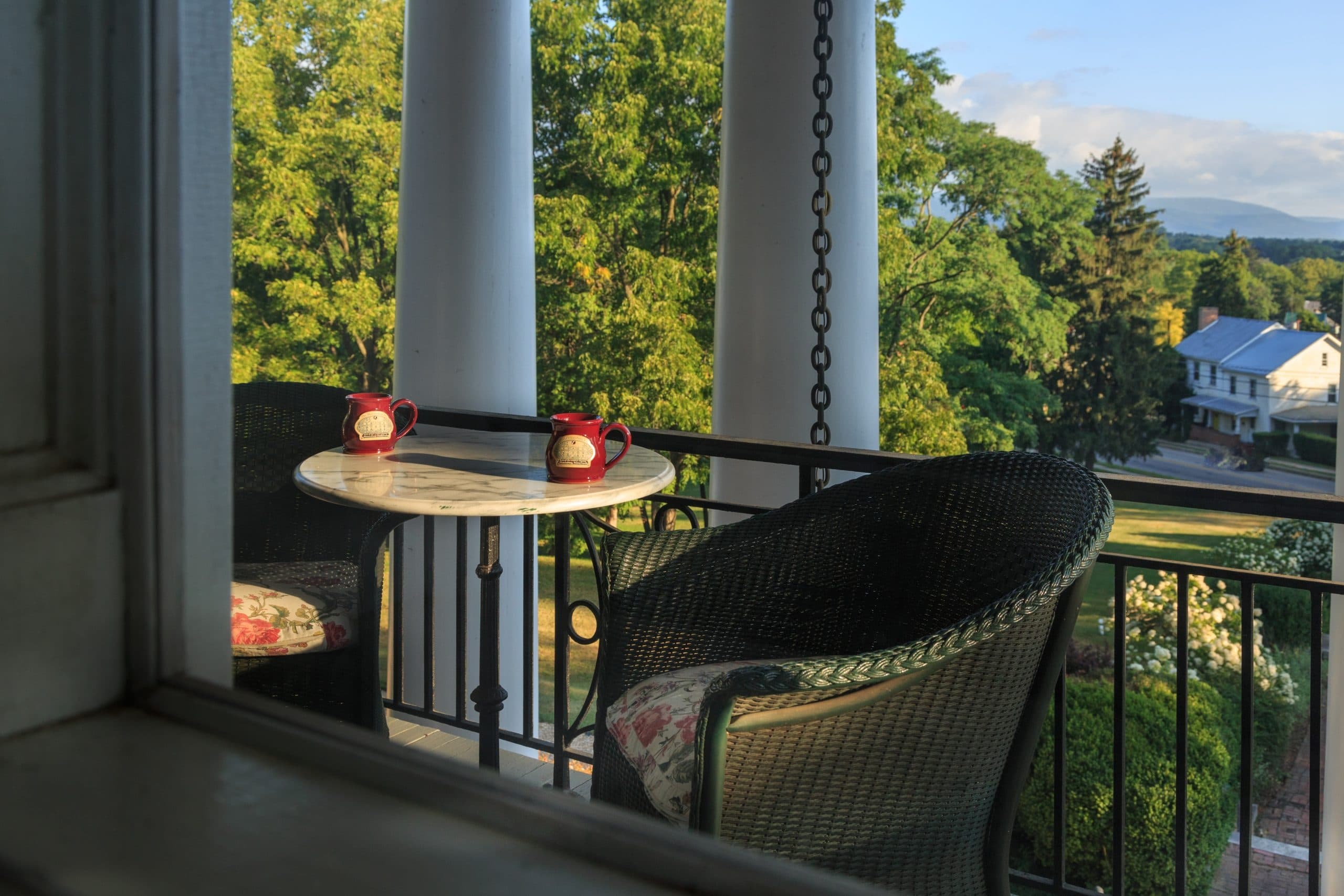 A cozy balcony with a small table, two mugs, and a view of green trees and a distant house.