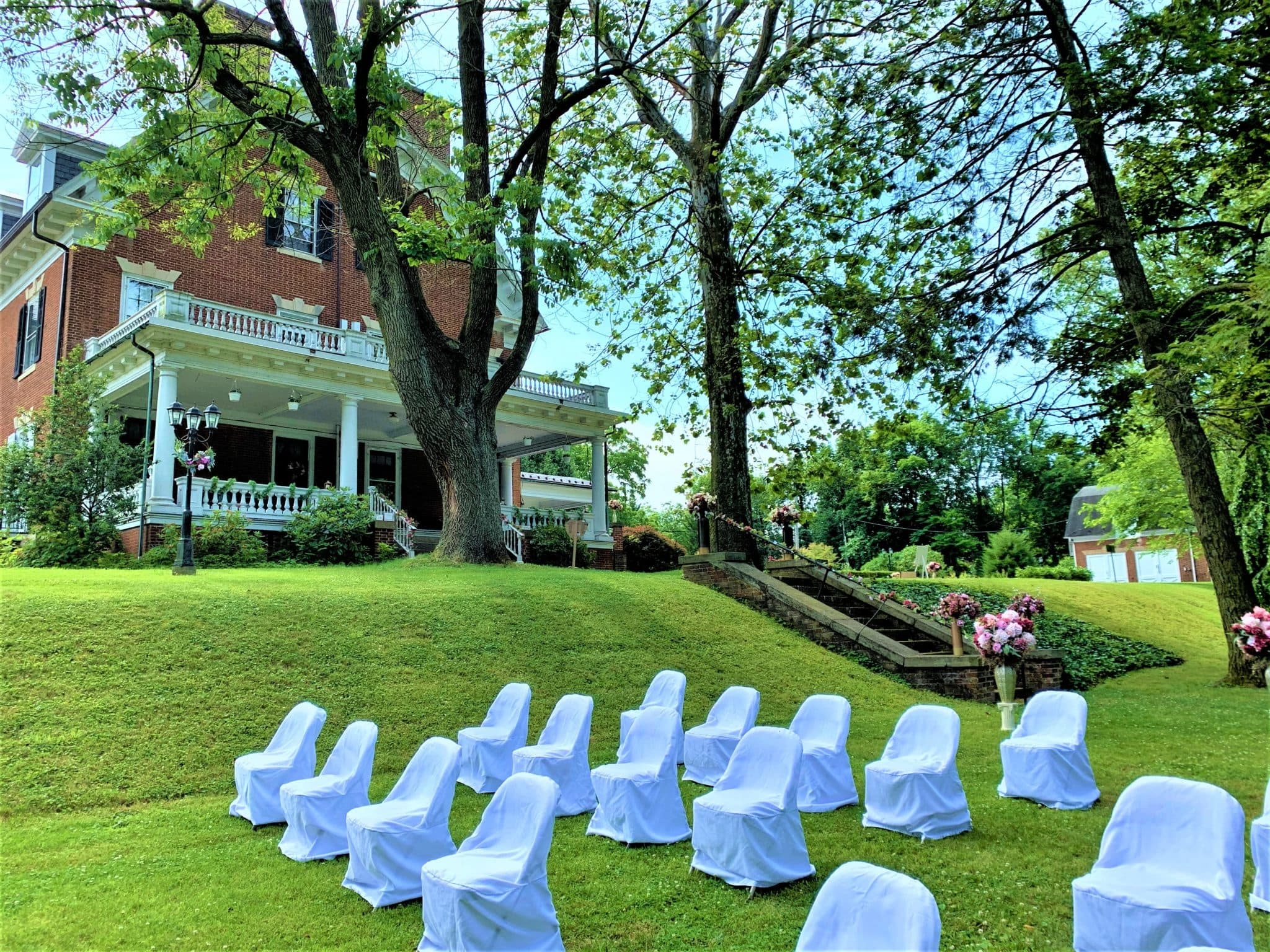 A beautifully landscaped garden with white chairs arranged for an outdoor event in front of a historic house.