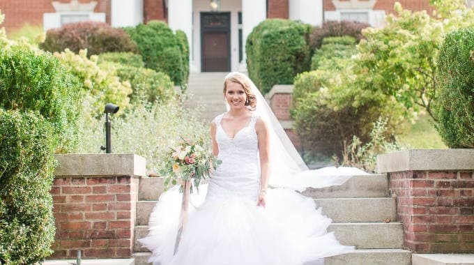 A smiling bride in a flowing white gown descends the steps of a venue, holding a bouquet amidst lush greenery.