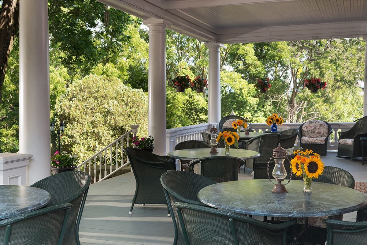 Covered porch with round tables and wicker chairs, decorated with sunflower bouquets. Green trees and blooming plants create a serene, inviting atmosphere. Covered porch with round tables and wicker chairs, decorated with sunflower bouquets. Green trees and blooming plants create a serene, inviting atmosphere.