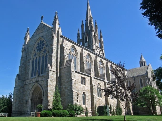 Gothic-style stone church with tall spires and arched windows against a clear blue sky. Green trees and shrubs surround the building, conveying serenity. Gothic-style stone church with tall spires and arched windows against a clear blue sky. Green trees and shrubs surround the building, conveying serenity.