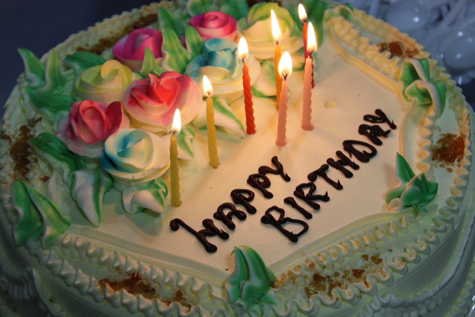A birthday cake with "Happy Birthday" written in black icing. Five lit candles surrounded by colorful icing roses create a festive, joyful mood.