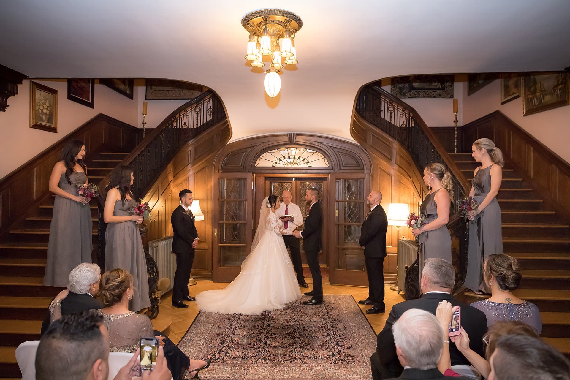 A bride and groom stand before an officiant in an elegant hall with wooden staircases. Bridesmaids in gray dresses and groomsmen flank the couple. Guests watch seated. A bride and groom stand before an officiant in an elegant hall with wooden staircases. Bridesmaids in gray dresses and groomsmen flank the couple. Guests watch seated.