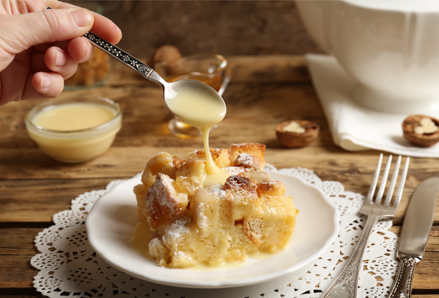 A hand holds a spoon pouring creamy sauce over a slice of bread pudding on a lace-trimmed plate, with nuts and sauce-filled bowls in the background. A hand holds a spoon pouring creamy sauce over a slice of bread pudding on a lace-trimmed plate, with nuts and sauce-filled bowls in the background.