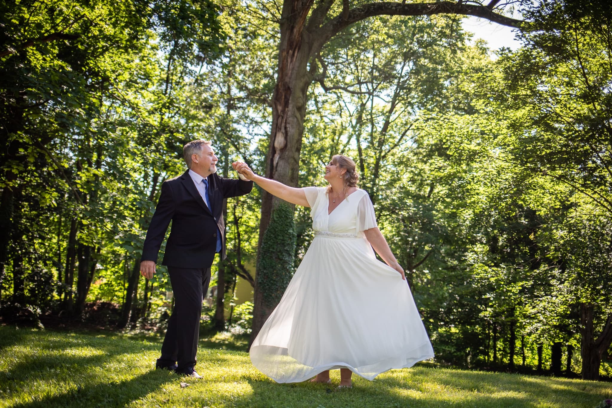A couple dances joyfully on a sunlit lawn surrounded by trees. The woman twirls in a flowing white dress, holding the man's hand as he smiles in a dark suit. A couple dances joyfully on a sunlit lawn surrounded by trees. The woman twirls in a flowing white dress, holding the man's hand as he smiles in a dark suit.