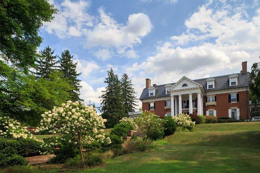 Historic brick mansion with white columns, surrounded by lush green lawn and blooming bushes under a partly cloudy blue sky, evoking elegance and tranquility. Historic brick mansion with white columns, surrounded by lush green lawn and blooming bushes under a partly cloudy blue sky, evoking elegance and tranquility.