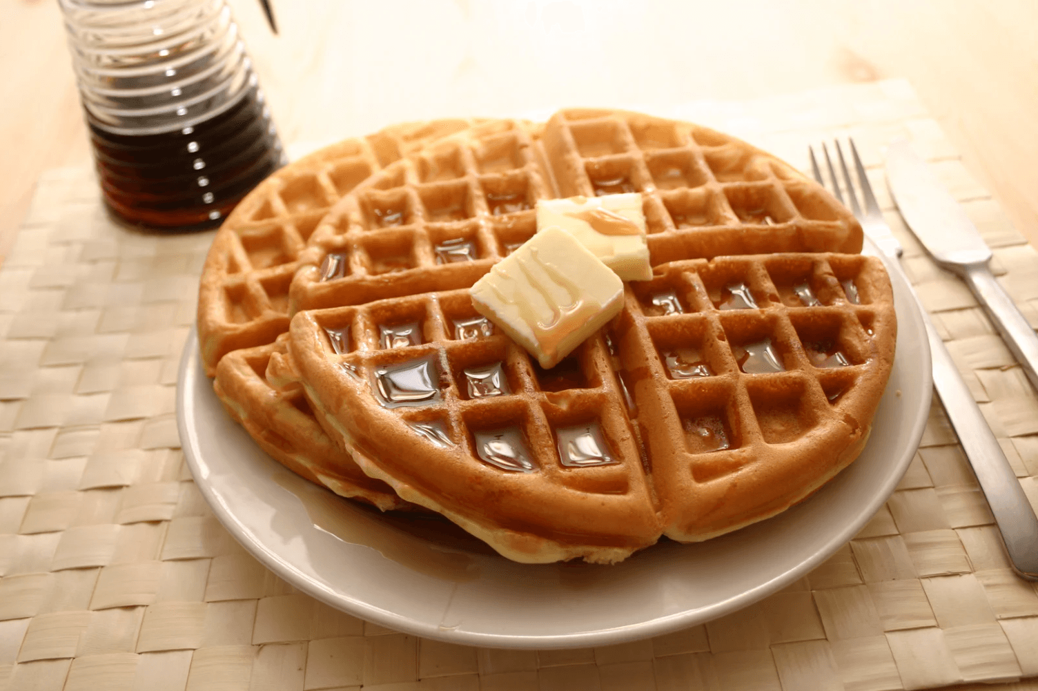 A plate of golden waffles topped with melting butter and syrup, next to a bottle of syrup and a fork and knife on a woven placemat. Warm and inviting. A plate of golden waffles topped with melting butter and syrup, next to a bottle of syrup and a fork and knife on a woven placemat. Warm and inviting.