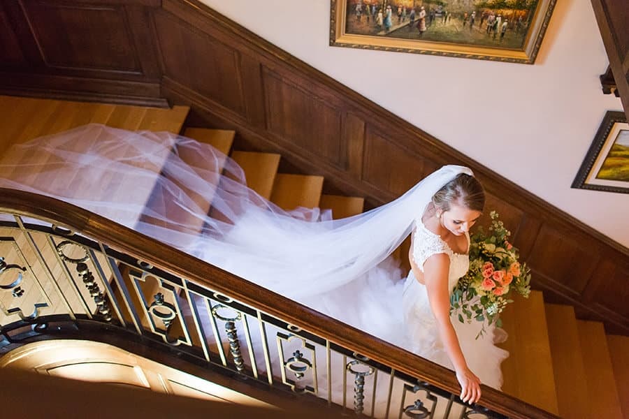 Bride descends wooden staircase, holding a bouquet of peach and white flowers. Her long veil trails behind, creating an elegant, serene atmosphere.