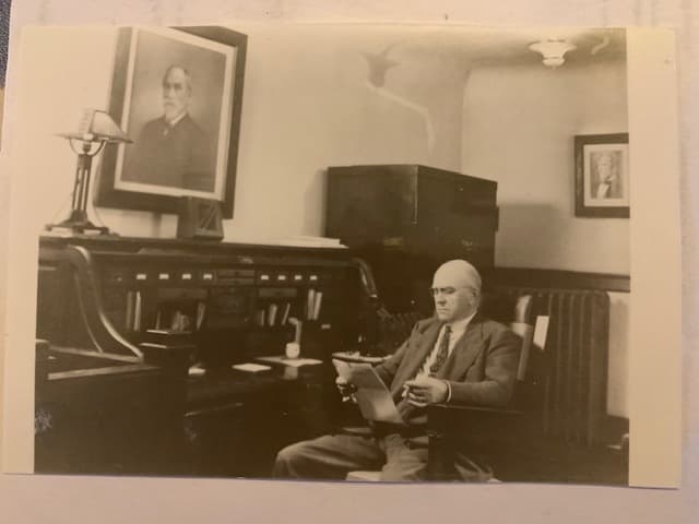 Black and white photo of a man in a suit sitting at a desk in a vintage office. He reads papers, with framed portraits on the walls, evoking a historical atmosphere. Black and white photo of a man in a suit sitting at a desk in a vintage office. He reads papers, with framed portraits on the walls, evoking a historical atmosphere.