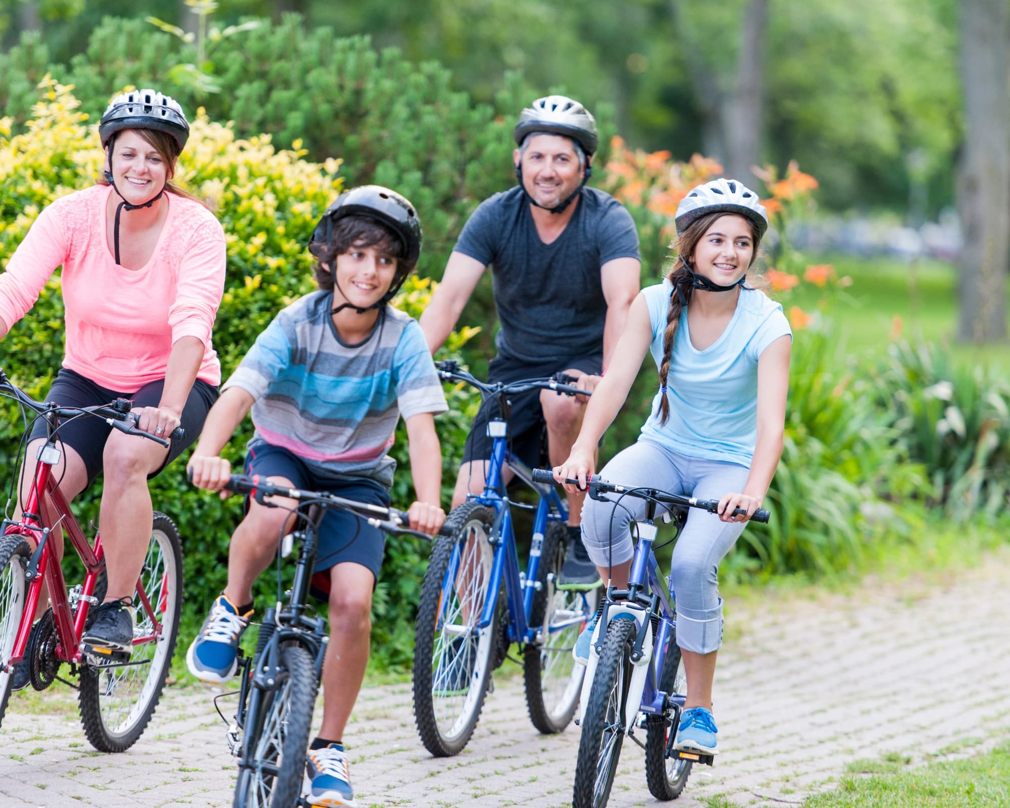 A family of four, wearing helmets, smiles while riding bikes on a park path. Green trees and colorful flowers create a cheerful, sunny backdrop. A family of four, wearing helmets, smiles while riding bikes on a park path. Green trees and colorful flowers create a cheerful, sunny backdrop.