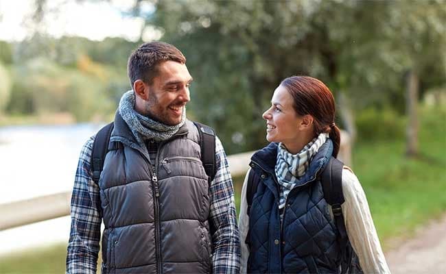 A smiling couple wearing vests and scarves walks along a wooded path. The background is blurred, suggesting a peaceful, outdoor setting.