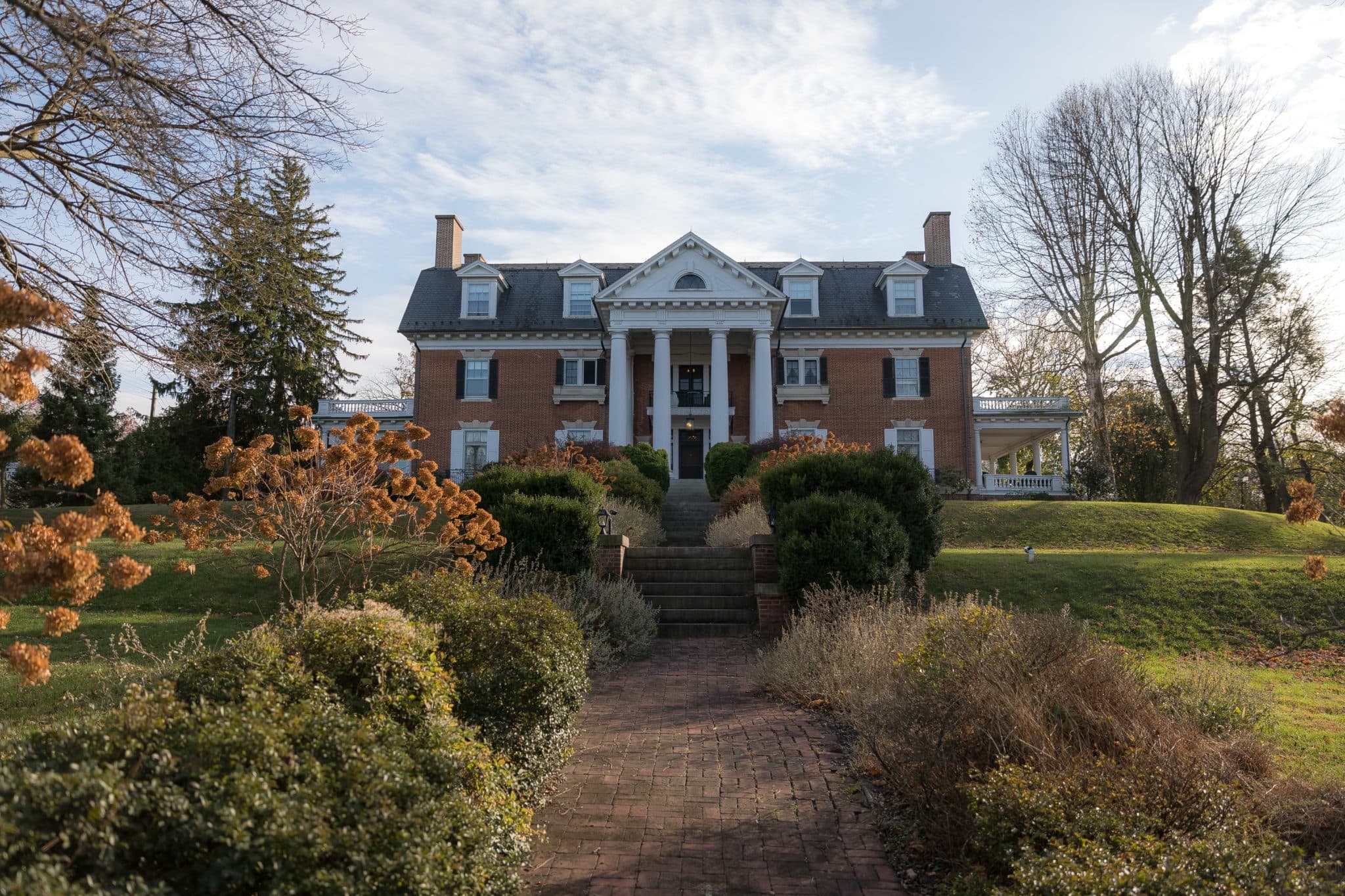 A grand brick mansion with white pillars and a slate roof stands amid a landscaped garden. Bare trees and autumn bushes under a partly cloudy sky. A grand brick mansion with white pillars and a slate roof stands amid a landscaped garden. Bare trees and autumn bushes under a partly cloudy sky.
