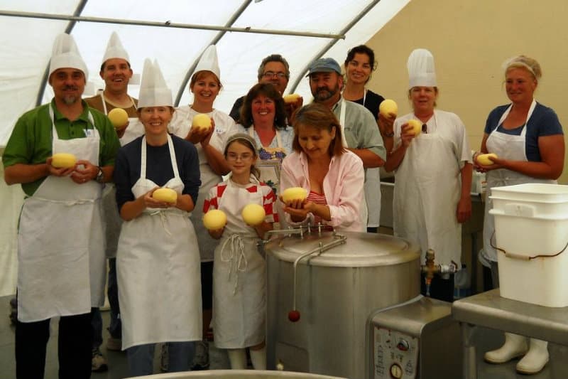 Group of twelve smiling people, some in chef hats, hold yellow balls of cheese in a bright dairy kitchen, exuding joy and teamwork Group of twelve smiling people, some in chef hats, hold yellow balls of cheese in a bright dairy kitchen, exuding joy and teamwork