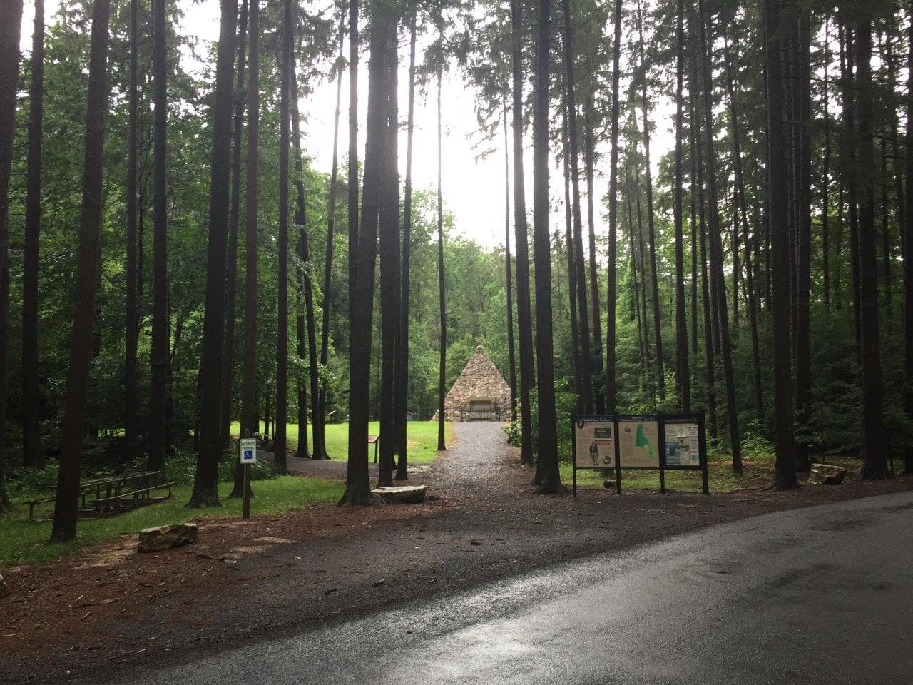 A stone pavilion sits in a clearing surrounded by tall trees. Informational signs and a picnic table are visible near a paved road in the foreground. A stone pavilion sits in a clearing surrounded by tall trees. Informational signs and a picnic table are visible near a paved road in the foreground.