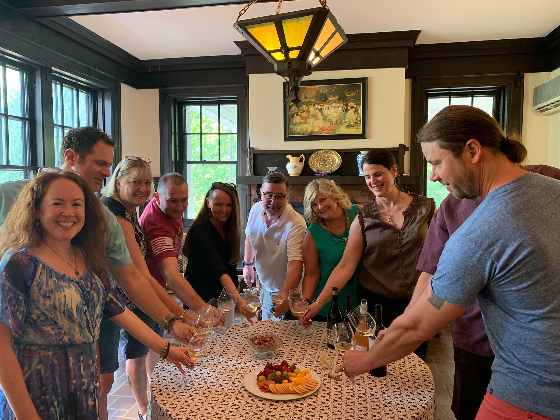 A group of nine people stands around a table with wine glasses raised in a toast, smiling. The table is set with a lace cloth, cheese, and fruit, conveying a joyful atmosphere. A group of nine people stands around a table with wine glasses raised in a toast, smiling. The table is set with a lace cloth, cheese, and fruit, conveying a joyful atmosphere.