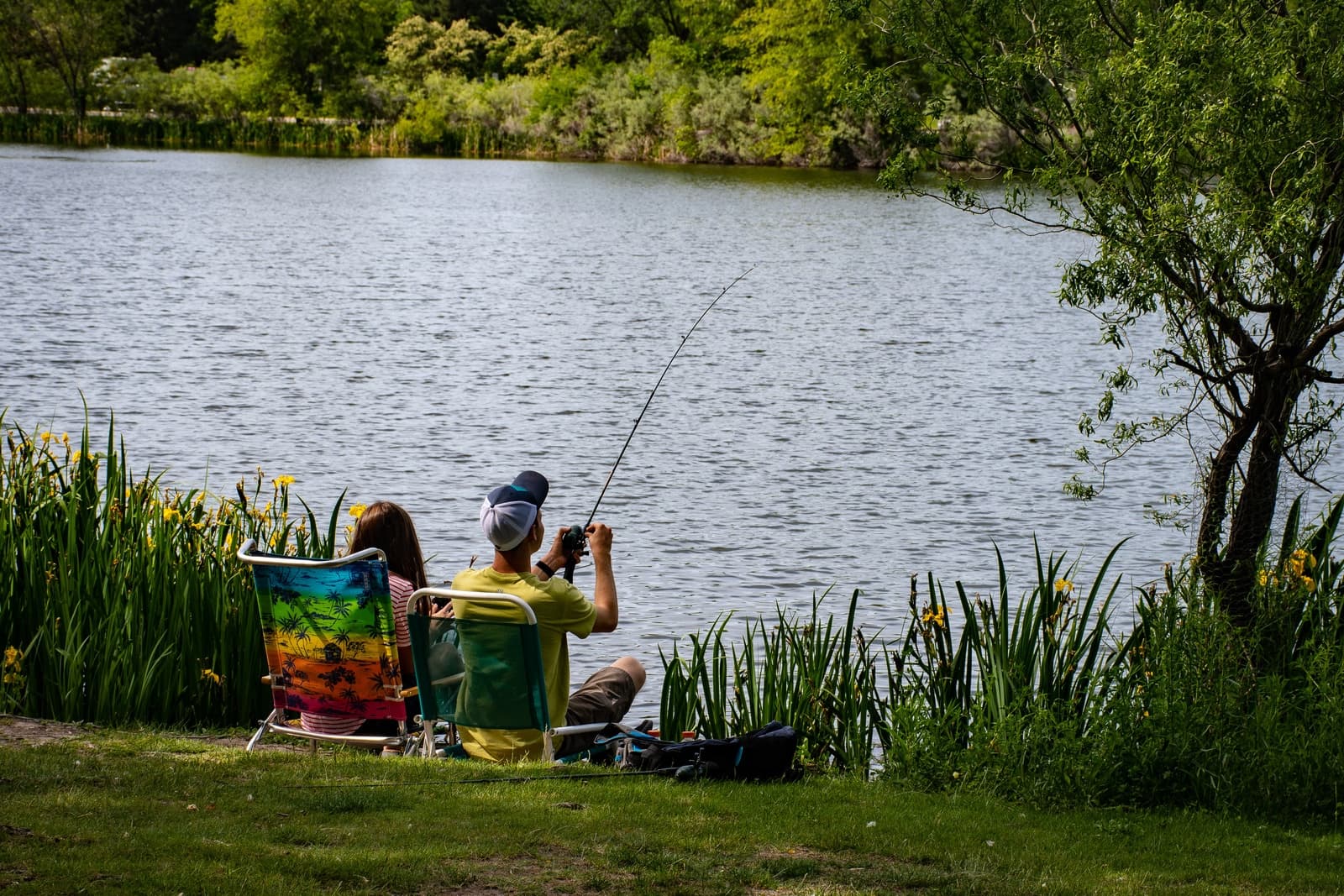 A couple sits on colorful chairs by a lake, fishing amidst lush greenery. The tranquil scene conveys relaxation and enjoyment of nature. A couple sits on colorful chairs by a lake, fishing amidst lush greenery. The tranquil scene conveys relaxation and enjoyment of nature.
