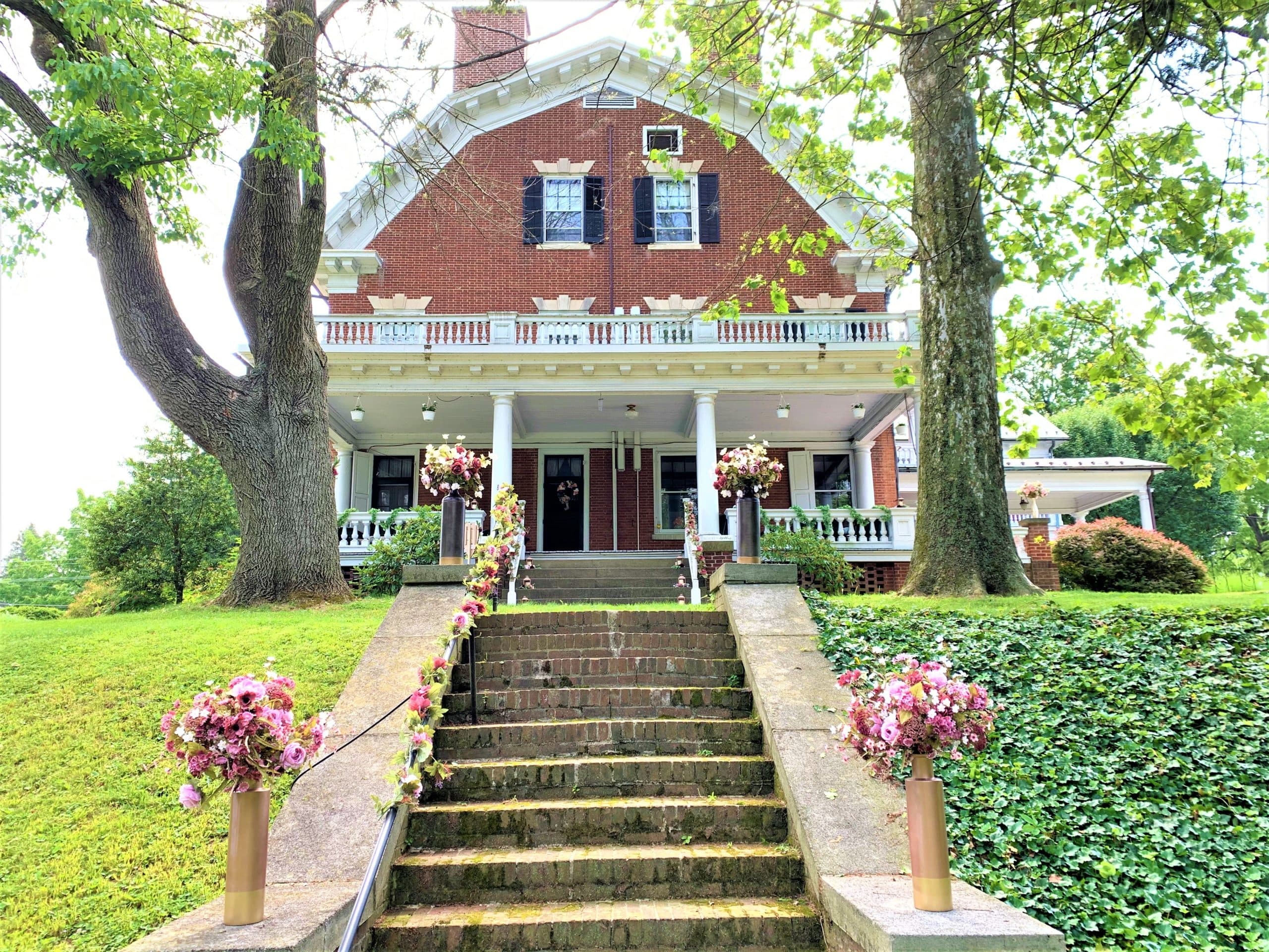 Historic brick house with black shutters, viewed from stone steps adorned with pink flowers. Lush green trees flank the inviting entrance. Historic brick house with black shutters, viewed from stone steps adorned with pink flowers. Lush green trees flank the inviting entrance.