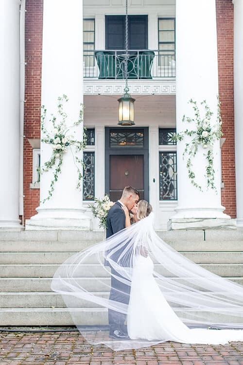 A bride and groom embrace on stone steps in front of a building with large white columns. The bride's veil flows dramatically in the foreground. A bride and groom embrace on stone steps in front of a building with large white columns. The bride's veil flows dramatically in the foreground.