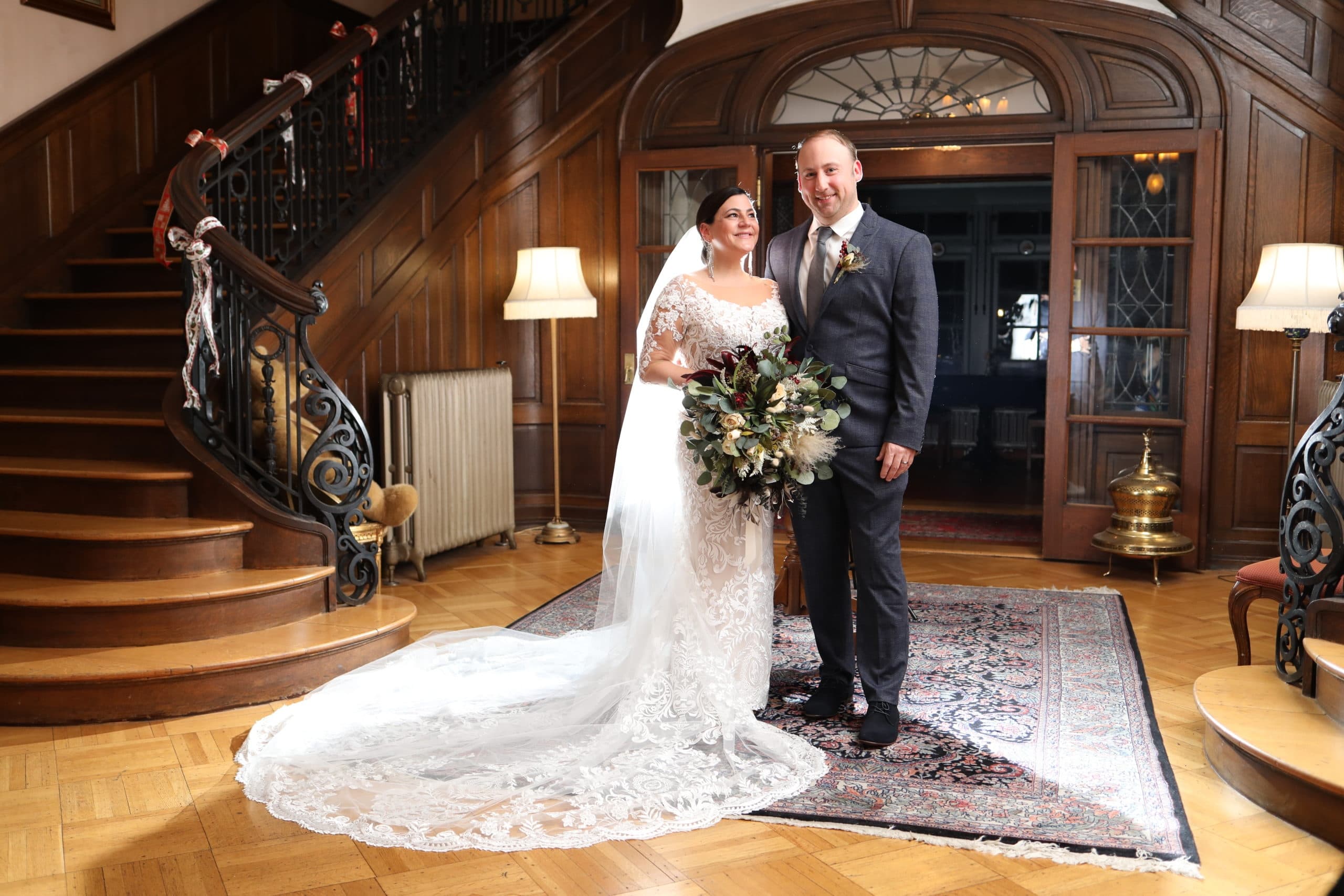 Bride in a lace gown and groom in a suit smile in a vintage room. They stand on a patterned rug by a wooden staircase, radiating joy. Bride in a lace gown and groom in a suit smile in a vintage room. They stand on a patterned rug by a wooden staircase, radiating joy.