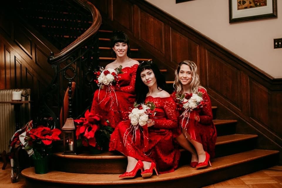 Three women in matching red dresses and shoes sit on a wooden staircase, holding bouquets of white flowers. They are surrounded by festive poinsettias, creating a joyful, celebratory atmosphere. Three women in matching red dresses and shoes sit on a wooden staircase, holding bouquets of white flowers. They are surrounded by festive poinsettias, creating a joyful, celebratory atmosphere.