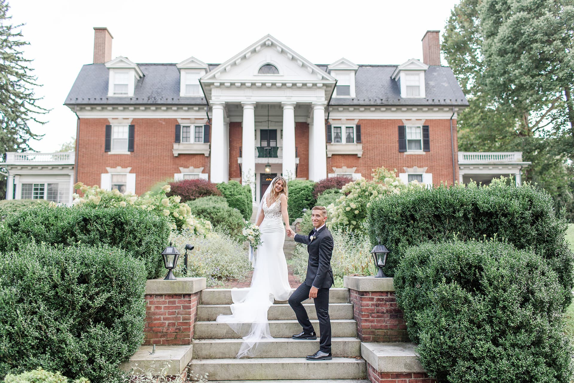Bride in white gown and groom in black suit stand on stone steps, hand in hand, in front of a grand brick mansion with white columns. Romantic setting. Bride in white gown and groom in black suit stand on stone steps, hand in hand, in front of a grand brick mansion with white columns. Romantic setting.