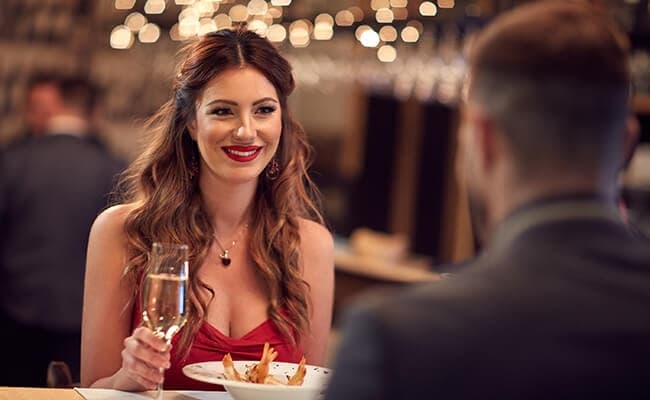 A woman in a red dress smiles, holding a glass of champagne at a dimly lit restaurant with bokeh lights. She is sitting across from a man. Romantic ambiance. A woman in a red dress smiles, holding a glass of champagne at a dimly lit restaurant with bokeh lights. She is sitting across from a man. Romantic ambiance.