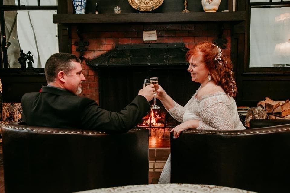 A couple sits in front of a lit fireplace, clinking champagne glasses. They share a joyful moment, smiling in a warm, cozy room with elegant decor. A couple sits in front of a lit fireplace, clinking champagne glasses. They share a joyful moment, smiling in a warm, cozy room with elegant decor.